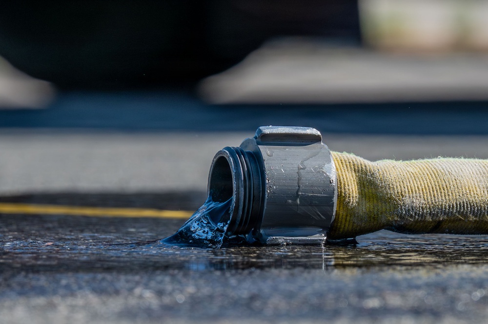 A close-up photograph of a fire hose coupling lying on wet asphalt, with water actively flowing out of the open metal connector end. The hose appears to be a standard yellow/olive-colored woven fire hose with a dark metal storz or threaded coupling. The image is taken at ground level with shallow depth of field, creating a sharp foreground and blurred background.