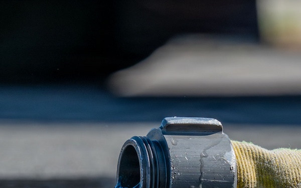 A close-up photograph of a fire hose coupling lying on wet asphalt, with water actively flowing out of the open metal connector end. The hose appears to be a standard yellow/olive-colored woven fire hose with a dark metal storz or threaded coupling. The image is taken at ground level with shallow depth of field, creating a sharp foreground and blurred background.