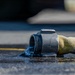 A close-up photograph of a fire hose coupling lying on wet asphalt, with water actively flowing out of the open metal connector end. The hose appears to be a standard yellow/olive-colored woven fire hose with a dark metal storz or threaded coupling. The image is taken at ground level with shallow depth of field, creating a sharp foreground and blurred background.