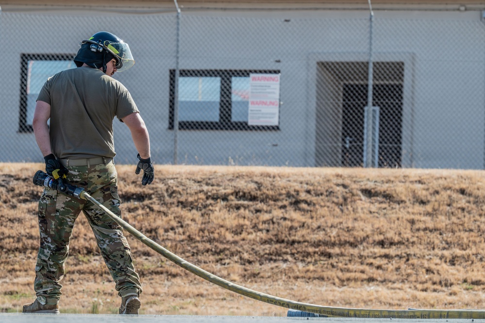 A military or emergency services personnel in multicam/OCP camouflage uniform is handling a fire hose during what appears to be a training exercise. The individual is wearing a black protective helmet with a face shield and tactical gloves. The setting appears to be a military installation or training facility with a chain-link fenced building and warning signs visible in the background.