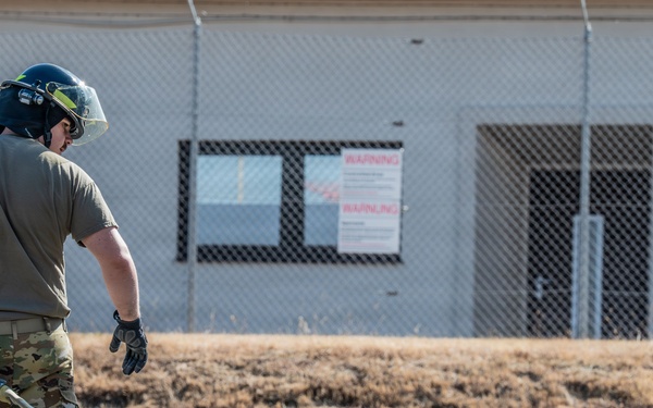 A military or emergency services personnel in multicam/OCP camouflage uniform is handling a fire hose during what appears to be a training exercise. The individual is wearing a black protective helmet with a face shield and tactical gloves. The setting appears to be a military installation or training facility with a chain-link fenced building and warning signs visible in the background.