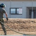 A military or emergency services personnel in multicam/OCP camouflage uniform is handling a fire hose during what appears to be a training exercise. The individual is wearing a black protective helmet with a face shield and tactical gloves. The setting appears to be a military installation or training facility with a chain-link fenced building and warning signs visible in the background.