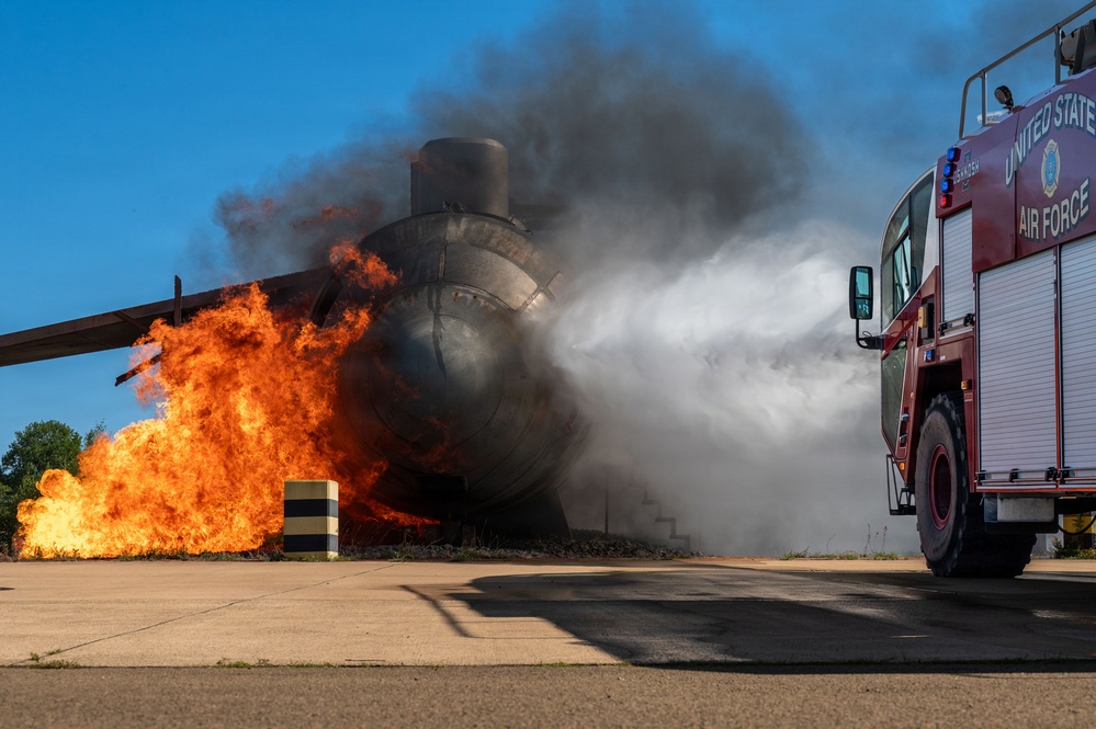 A United States Air Force fire truck is actively discharging a suppression agent (foam or water) onto a large fire engulfing what appears to be a mock aircraft fuselage or training prop on a paved surface. The scene depicts a firefighting training exercise, with intense orange flames and thick black smoke visible on the left side of the burning structure. The clear blue sky and controlled setting suggest this is a deliberate, supervised emergency response drill.