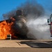 A United States Air Force fire truck is actively discharging a suppression agent (foam or water) onto a large fire engulfing what appears to be a mock aircraft fuselage or training prop on a paved surface. The scene depicts a firefighting training exercise, with intense orange flames and thick black smoke visible on the left side of the burning structure. The clear blue sky and controlled setting suggest this is a deliberate, supervised emergency response drill.