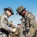 Two military personnel in different camouflage uniforms are engaged in a close interaction, appearing to inspect or adjust rappelling/climbing harness equipment. The soldier on the left wears what appears to be a Korean military uniform with Hangul script patches, while the soldier on the right wears USMC MARPAT digital camouflage. A group of additional military personnel in similar gear are visible in the background, suggesting a joint training exercise.