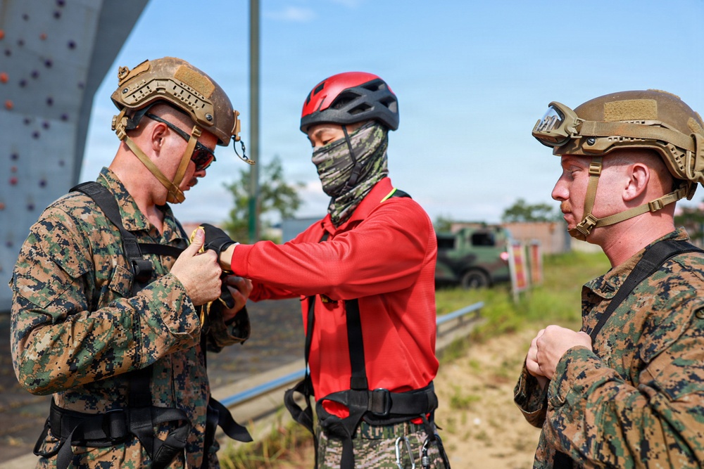 Three individuals are gathered outdoors near what appears to be a military training facility with a climbing wall visible in the background. Two individuals wearing U.S. Marine Corps MARPAT digital camouflage uniforms and tan tactical helmets are interacting with a third person wearing a red long-sleeve shirt, climbing harness, red helmet, and camouflage face covering. The central figure appears to be having equipment or gear checked/adjusted by one of the Marines.