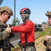 Three individuals are gathered outdoors near what appears to be a military training facility with a climbing wall visible in the background. Two individuals wearing U.S. Marine Corps MARPAT digital camouflage uniforms and tan tactical helmets are interacting with a third person wearing a red long-sleeve shirt, climbing harness, red helmet, and camouflage face covering. The central figure appears to be having equipment or gear checked/adjusted by one of the Marines.