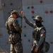Two military personnel are standing in front of a climbing wall, engaged in what appears to be rappelling or rope training. One soldier wearing MARPAT digital camouflage and a tactical helmet is being assisted or instructed by a second soldier in a different camouflage pattern who is holding a rope overhead. The setting suggests a military training facility with a multi-colored rock climbing wall in the background.