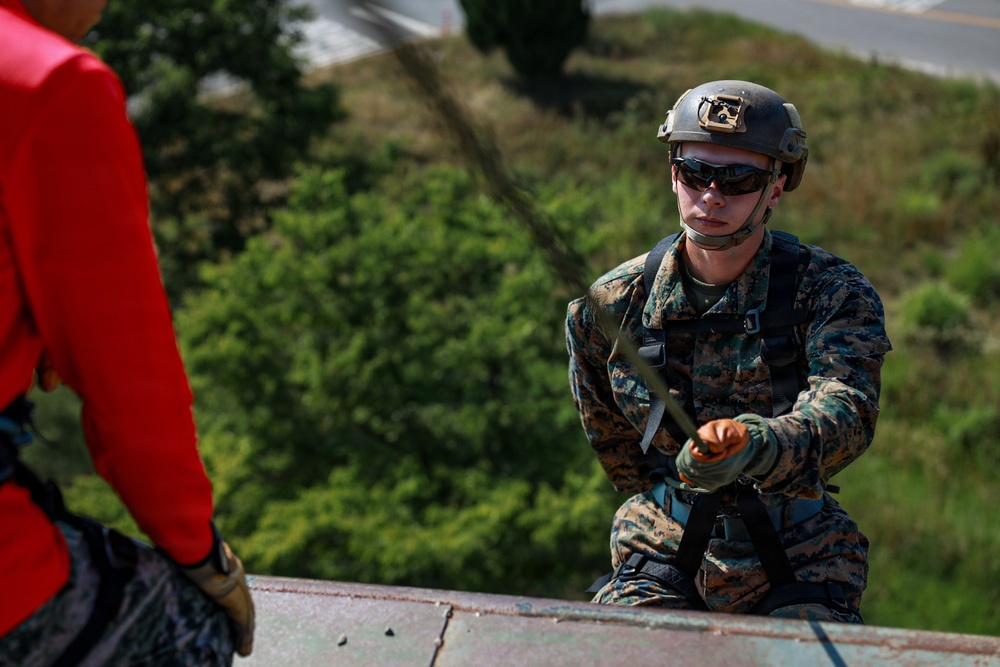 A U.S. Marine in full combat gear, including MARPAT camouflage uniform and tactical helmet, is positioned at the edge of an elevated structure, appearing to manage rappelling or climbing equipment. A second individual in a red shirt is partially visible on the left, also equipped with a harness. The setting appears to be an outdoor training exercise at a military installation.