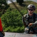 A U.S. Marine in full combat gear, including MARPAT camouflage uniform and tactical helmet, is positioned at the edge of an elevated structure, appearing to manage rappelling or climbing equipment. A second individual in a red shirt is partially visible on the left, also equipped with a harness. The setting appears to be an outdoor training exercise at a military installation.