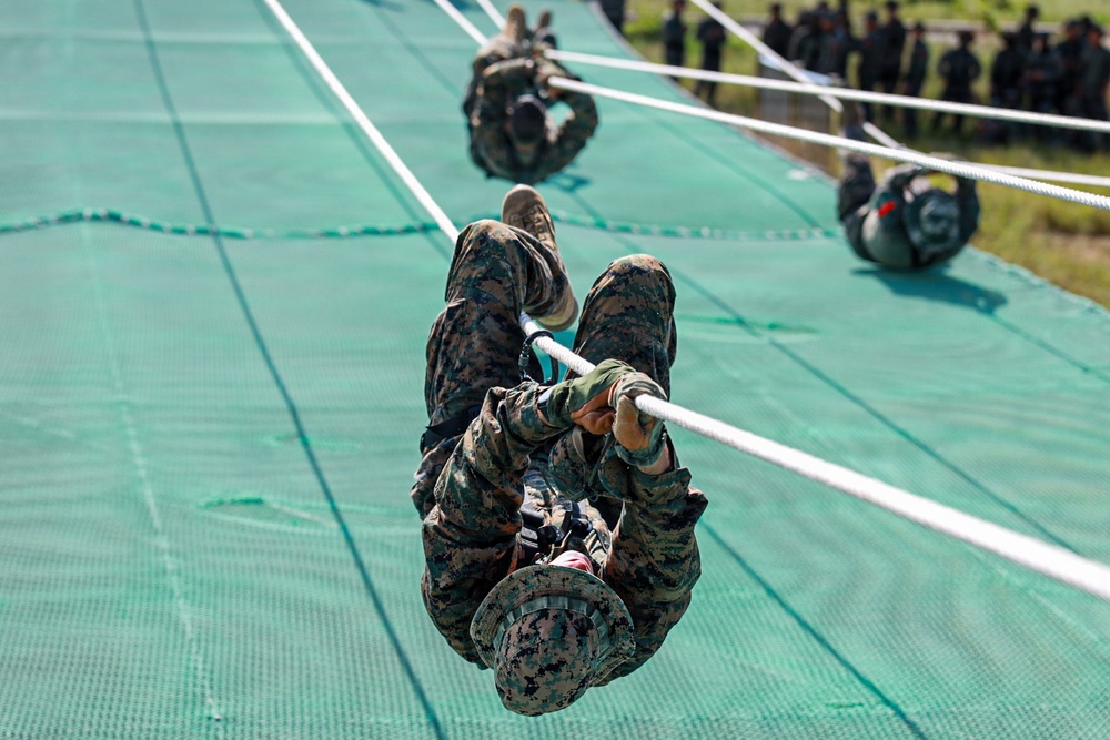 Military personnel in camouflage uniforms are traversing a rope obstacle course on a large green net surface. Multiple individuals are visible climbing or sliding along horizontal ropes in what appears to be a structured military training exercise. A group of observers in uniform can be seen watching from the background on the right side.