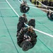 Military personnel in camouflage uniforms are traversing a rope obstacle course on a large green net surface. Multiple individuals are visible climbing or sliding along horizontal ropes in what appears to be a structured military training exercise. A group of observers in uniform can be seen watching from the background on the right side.