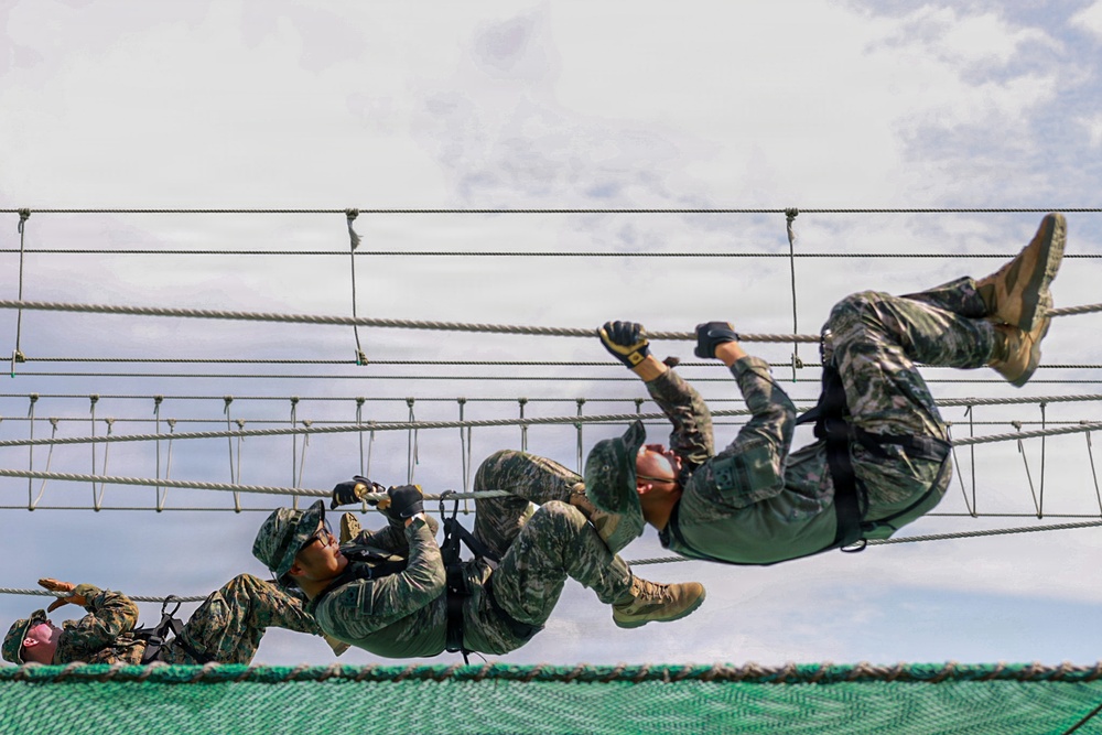 Multiple military personnel in camouflage uniforms are traversing a rope obstacle course suspended in the air. The soldiers are using hand-over-hand techniques to move along horizontal ropes while hanging beneath them. A safety net is visible below, indicating this is a controlled training exercise.