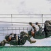 Multiple military personnel in camouflage uniforms are traversing a rope obstacle course suspended in the air. The soldiers are using hand-over-hand techniques to move along horizontal ropes while hanging beneath them. A safety net is visible below, indicating this is a controlled training exercise.