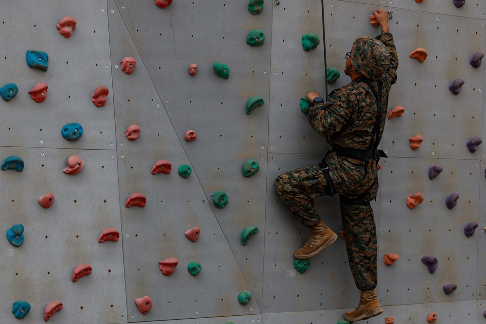 A person wearing military camouflage uniform and tan combat boots is climbing an indoor rock climbing wall equipped with colorful holds. The climber is secured with a harness and rope system, positioned on the right side of the wall mid-climb. The wall features multiple colored holds in pink, teal, blue, and purple arranged across a gray metal panel surface.