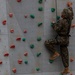 A person wearing military camouflage uniform and tan combat boots is climbing an indoor rock climbing wall equipped with colorful holds. The climber is secured with a harness and rope system, positioned on the right side of the wall mid-climb. The wall features multiple colored holds in pink, teal, blue, and purple arranged across a gray metal panel surface.