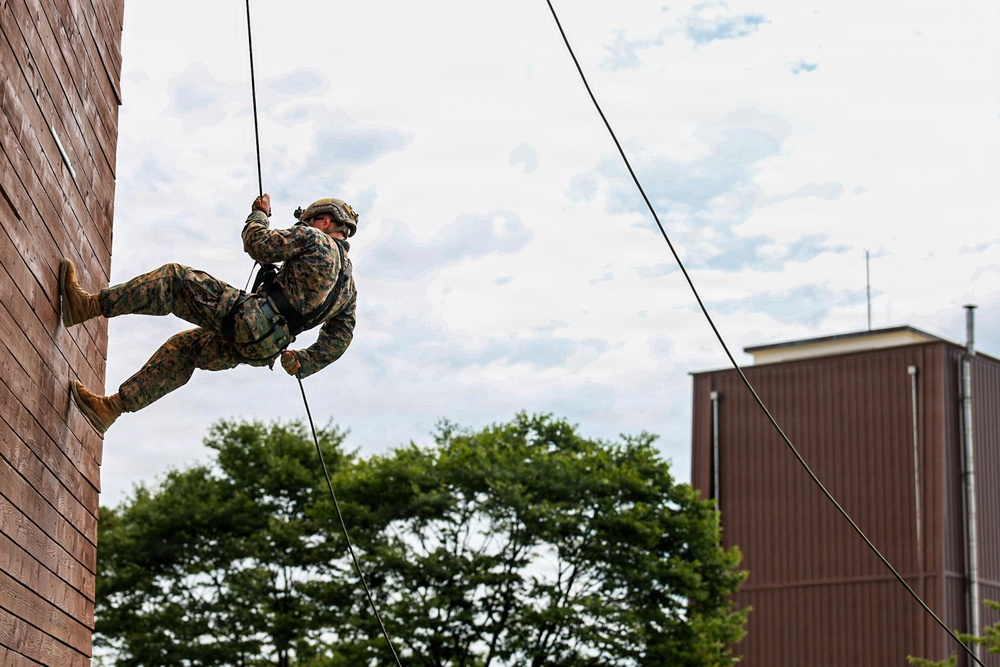 A military service member in camouflage uniform and helmet is rappelling down a wooden training wall. The individual is using ropes and a harness system in what appears to be a controlled training exercise at a military installation. Trees and a brown industrial or institutional building are visible in the background.