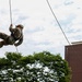 A military service member in camouflage uniform and helmet is rappelling down a wooden training wall. The individual is using ropes and a harness system in what appears to be a controlled training exercise at a military installation. Trees and a brown industrial or institutional building are visible in the background.