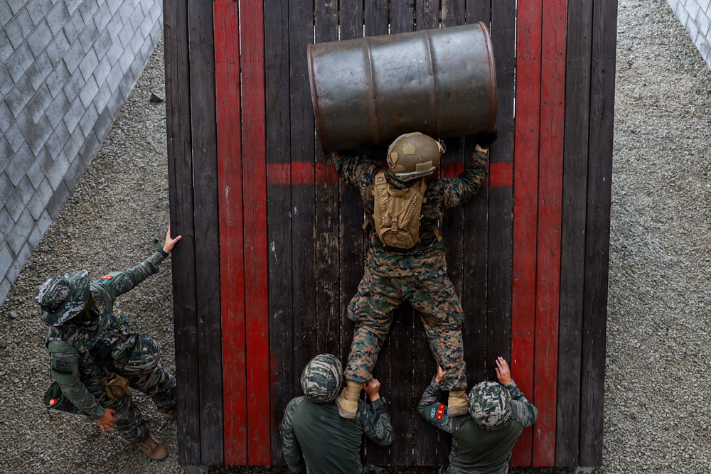 An aerial view shows a military training exercise where one soldier in US Marine Corps digital camouflage is scaling a tall wooden wall obstacle while carrying a large metal barrel on their back. Three other soldiers in different camouflage patterns are assisting from below by boosting and supporting the climber. The scene appears to be a team-based obstacle course challenge at a military training facility.