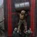 An aerial view shows a military training exercise where one soldier in US Marine Corps digital camouflage is scaling a tall wooden wall obstacle while carrying a large metal barrel on their back. Three other soldiers in different camouflage patterns are assisting from below by boosting and supporting the climber. The scene appears to be a team-based obstacle course challenge at a military training facility.