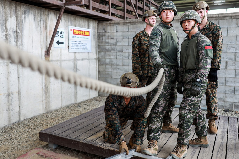 A group of military personnel in camouflage uniforms are gathered on a wooden platform near a thick rope, appearing to be engaged in a training exercise. The setting includes Korean-language signage on a concrete block wall, suggesting this is a joint military training facility in South Korea. Personnel wearing both US Marine Corps digital camouflage and Republic of Korea (ROK) Army camouflage patterns are visible, indicating a joint training exercise.