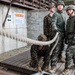 A group of military personnel in camouflage uniforms are gathered on a wooden platform near a thick rope, appearing to be engaged in a training exercise. The setting includes Korean-language signage on a concrete block wall, suggesting this is a joint military training facility in South Korea. Personnel wearing both US Marine Corps digital camouflage and Republic of Korea (ROK) Army camouflage patterns are visible, indicating a joint training exercise.
