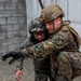 Two military personnel in camouflage uniforms are engaged in what appears to be a training exercise near a concrete block wall. One soldier wearing MARPAT (US Marine) pattern camouflage and a tan tactical helmet is assisting or restraining a second soldier wearing a different digital camouflage pattern and glasses. The interaction suggests a casualty evacuation drill or combatives training scenario.