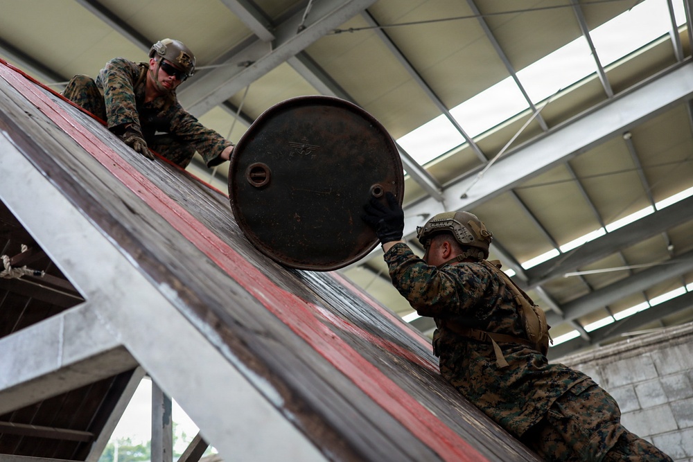 Two military personnel in camouflage uniforms are working together to move a large metal drum or barrel up an inclined ramp inside an industrial or warehouse-type structure. Both individuals are wearing combat helmets and military-pattern (MARPAT) uniforms consistent with U.S. Marines. The image is taken from a low angle, emphasizing the effort involved in lifting the heavy cylindrical object.