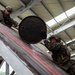 Two military personnel in camouflage uniforms are working together to move a large metal drum or barrel up an inclined ramp inside an industrial or warehouse-type structure. Both individuals are wearing combat helmets and military-pattern (MARPAT) uniforms consistent with U.S. Marines. The image is taken from a low angle, emphasizing the effort involved in lifting the heavy cylindrical object.