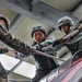 Military personnel from at least two different armed forces are engaged in a rope-based training exercise, likely a rappelling or obstacle course activity. Two soldiers wearing olive-green uniforms with helmets and red-accented patches are working with thick rope lines alongside personnel in MARPAT (US Marine) digital camouflage uniforms. The setting appears to be an indoor or covered training facility with visible steel structural beams overhead.