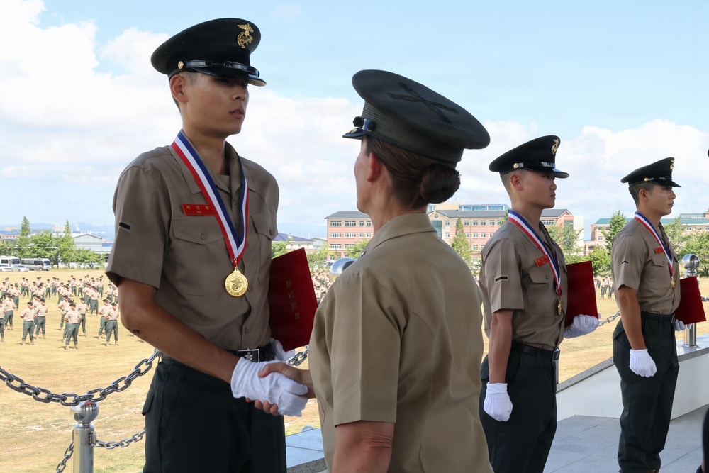 A military awards ceremony is taking place outdoors, likely at a Korean military installation. A senior officer in tan/khaki uniform is presenting medals and red certificates to uniformed recipients wearing gold medals on red-white-blue ribbons. A large formation of troops is visible in the background on a parade ground.