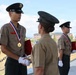 A military awards ceremony is taking place outdoors, likely at a Korean military installation. A senior officer in tan/khaki uniform is presenting medals and red certificates to uniformed recipients wearing gold medals on red-white-blue ribbons. A large formation of troops is visible in the background on a parade ground.