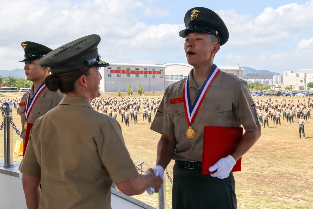 A military awards ceremony is taking place on an elevated platform, where a senior officer is shaking hands with a recipient wearing a gold medal on a red-white-and-blue ribbon lanyard. The recipient holds a red folder and wears white gloves, suggesting a formal recognition event. In the background, a large formation of uniformed personnel stands at attention on a parade ground.