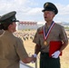 A military awards ceremony is taking place on an elevated platform, where a senior officer is shaking hands with a recipient wearing a gold medal on a red-white-and-blue ribbon lanyard. The recipient holds a red folder and wears white gloves, suggesting a formal recognition event. In the background, a large formation of uniformed personnel stands at attention on a parade ground.