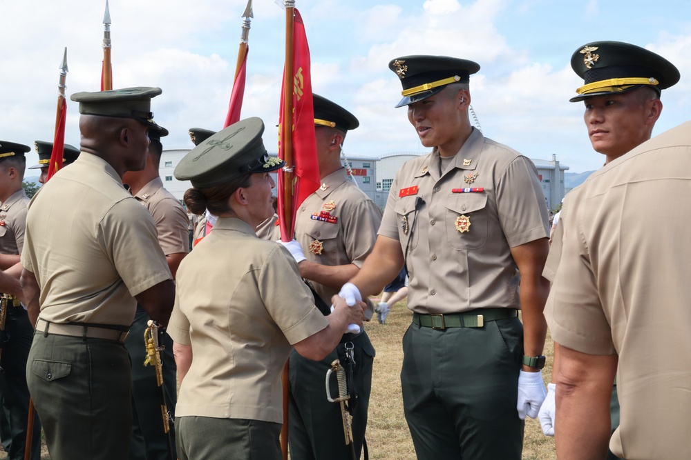 A military ceremony is taking place outdoors, likely a change of command or awards ceremony. A senior officer in a tan/khaki uniform with multiple medals is shaking hands with a female officer in olive green uniform. Red ceremonial flags/guidons are visible in the background alongside multiple uniformed personnel.
