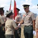 A military ceremony is taking place outdoors, likely a change of command or awards ceremony. A senior officer in a tan/khaki uniform with multiple medals is shaking hands with a female officer in olive green uniform. Red ceremonial flags/guidons are visible in the background alongside multiple uniformed personnel.