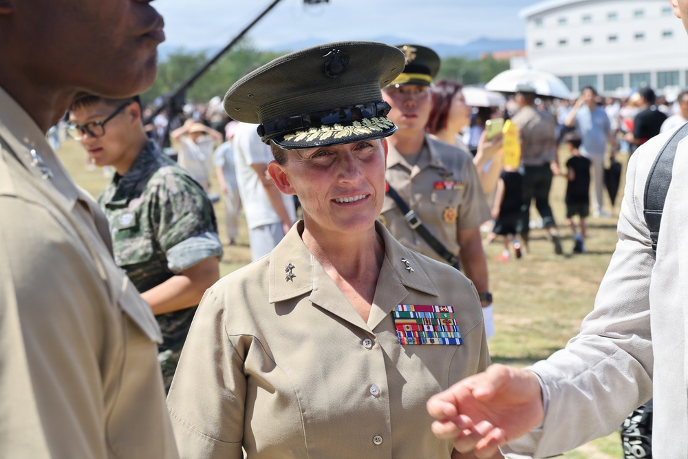 A female U.S. Marine Corps officer wearing a service uniform with two-star rank insignia (Major General) and multiple service ribbons is engaged in conversation at what appears to be an outdoor military event or air show. She is wearing the distinctive Marine Corps service cover with gold oak leaf embroidery indicating senior officer rank. The setting appears to be in an East Asian country, likely South Korea or Japan, based on the crowd and surrounding personnel.