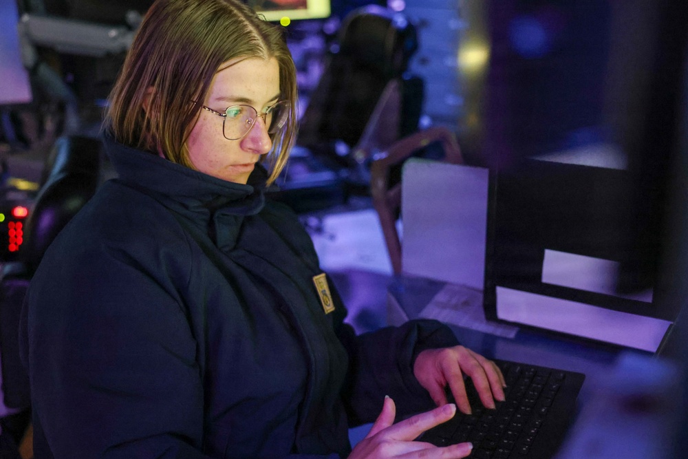 A young woman wearing glasses and a dark navy jacket is focused on typing at a computer keyboard in a dimly lit environment. The setting appears to be a technical operations room or command center, illuminated by blue and purple ambient lighting. Multiple monitors and equipment are visible in the background.