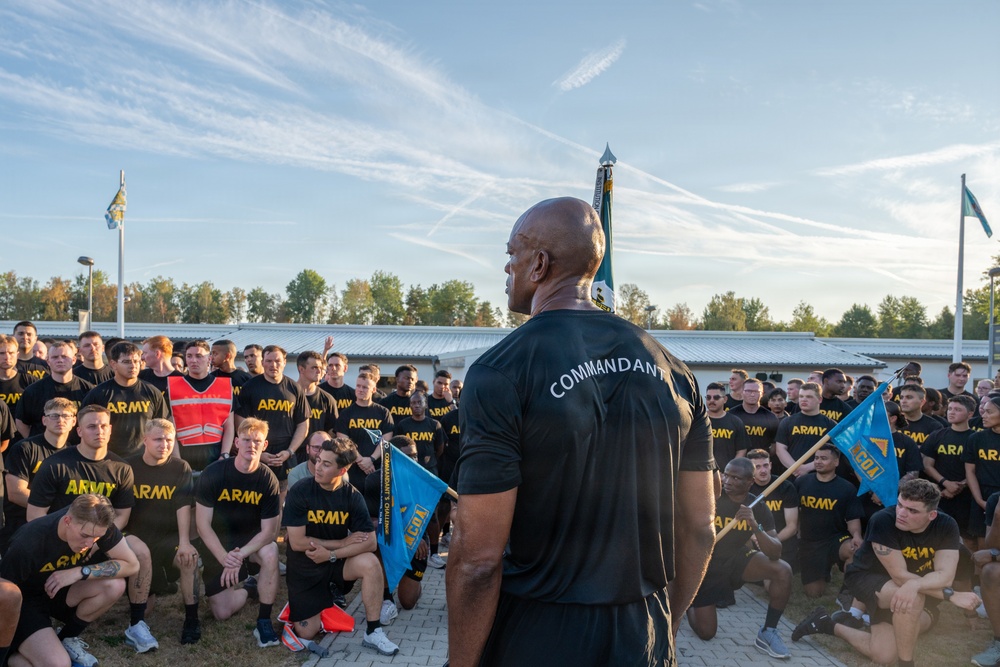 A person wearing a black shirt labeled 'COMMANDANT' addresses a large formation of U.S. Army soldiers, all dressed in black 'ARMY' physical training shirts. The gathering appears to be an outdoor military formation or briefing, likely before a physical training event or challenge. Several blue pennant flags labeled 'ACOA' are visible among the assembled soldiers.
