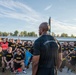 A person wearing a black shirt labeled 'COMMANDANT' addresses a large formation of U.S. Army soldiers, all dressed in black 'ARMY' physical training shirts. The gathering appears to be an outdoor military formation or briefing, likely before a physical training event or challenge. Several blue pennant flags labeled 'ACOA' are visible among the assembled soldiers.