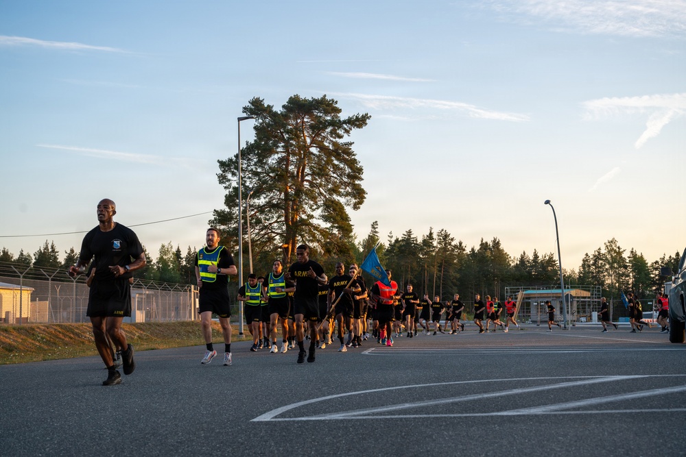 A large group of U.S. Army soldiers in black PT (physical training) uniforms are conducting a formation run on a paved road at what appears to be a military installation. Several participants wear high-visibility yellow safety vests, and one soldier carries a blue unit guidon flag. The scene is set during golden hour lighting, with tall pine trees and a fenced military compound visible in the background.