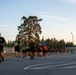 A large group of U.S. Army soldiers in black PT (physical training) uniforms are conducting a formation run on a paved road at what appears to be a military installation. Several participants wear high-visibility yellow safety vests, and one soldier carries a blue unit guidon flag. The scene is set during golden hour lighting, with tall pine trees and a fenced military compound visible in the background.