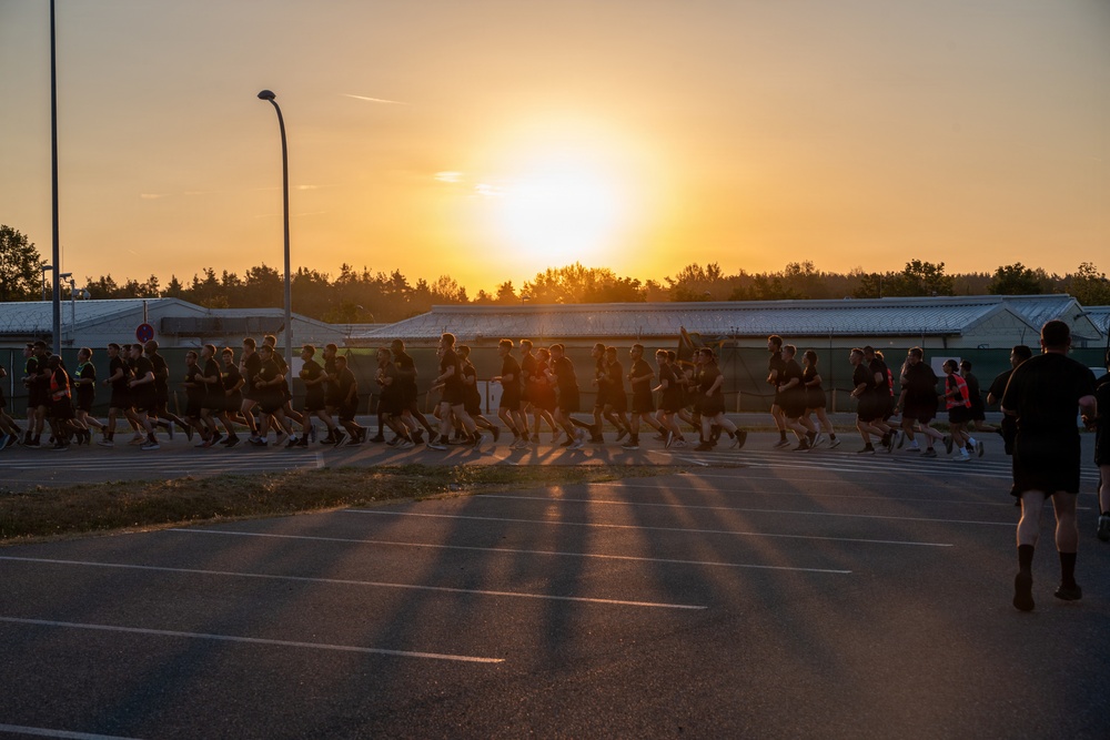 A large group of people dressed in dark athletic clothing are running together in formation across a parking lot during sunrise or sunset. The golden light from the low sun creates dramatic long shadows across the asphalt. The setting appears to be a military base or institutional facility, suggesting this is an organized physical training run.
