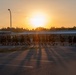 A large group of people dressed in dark athletic clothing are running together in formation across a parking lot during sunrise or sunset. The golden light from the low sun creates dramatic long shadows across the asphalt. The setting appears to be a military base or institutional facility, suggesting this is an organized physical training run.