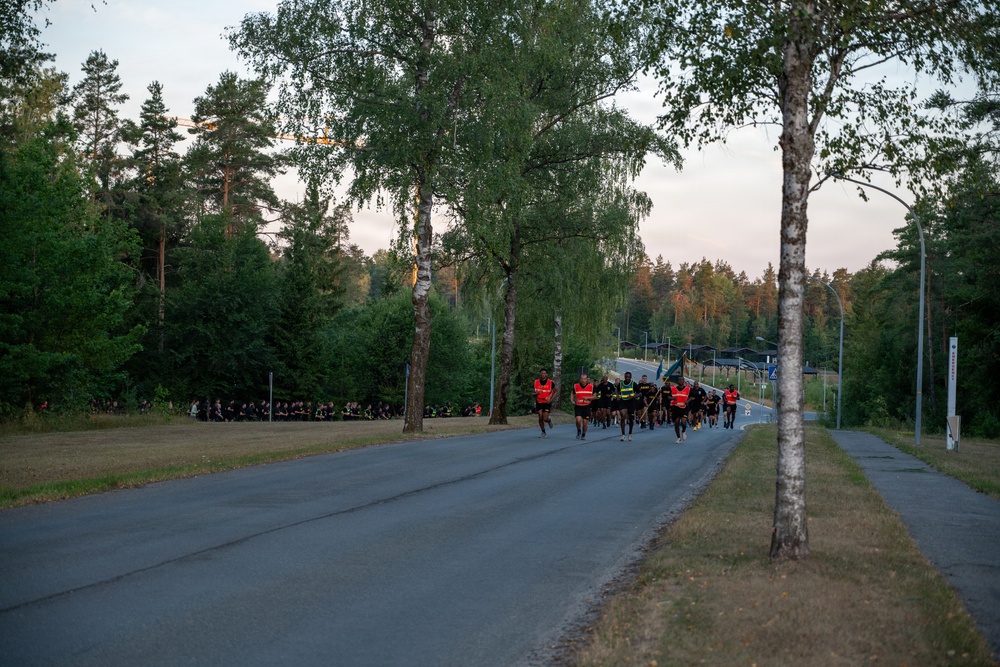 A group of runners is moving along a paved road through a forested area during early morning or evening light. Several participants are wearing high-visibility orange safety vests, suggesting an organized run or military physical training event. A large group of people dressed in dark clothing can be seen seated on the grass to the left, likely spectators or waiting participants.