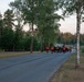 A group of runners is moving along a paved road through a forested area during early morning or evening light. Several participants are wearing high-visibility orange safety vests, suggesting an organized run or military physical training event. A large group of people dressed in dark clothing can be seen seated on the grass to the left, likely spectators or waiting participants.