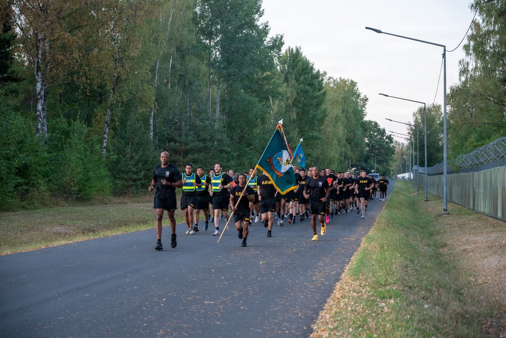 A large formation of U.S. Army soldiers in physical training (PT) uniforms — black shorts and shirts — are running in a group along a paved road. Several soldiers near the front are carrying military unit guidon flags, and some wear high-visibility yellow safety vests. The setting appears to be a military installation surrounded by trees, with a security fence and razor wire visible on the right side.
