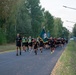 A large formation of U.S. Army soldiers in physical training (PT) uniforms — black shorts and shirts — are running in a group along a paved road. Several soldiers near the front are carrying military unit guidon flags, and some wear high-visibility yellow safety vests. The setting appears to be a military installation surrounded by trees, with a security fence and razor wire visible on the right side.