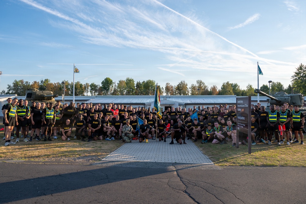 A large group of U.S. Army personnel pose for a formal group photograph at what appears to be a military installation, likely in Europe based on the signage and surroundings. The majority are wearing black Army PT (Physical Training) uniforms, with several individuals in yellow/green safety vests on the flanks. Military equipment including what appears to be a helicopter and artillery piece are visible in the background.