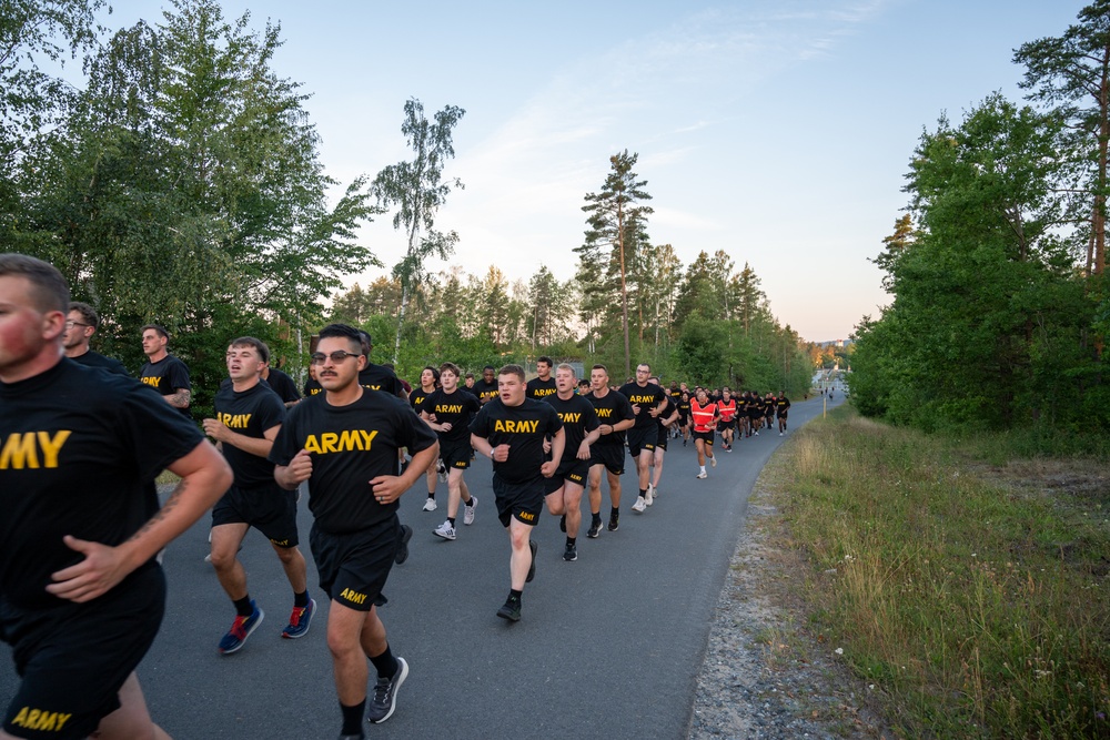 A large group of U.S. Army personnel are conducting a formation run along a paved road flanked by trees. All participants are wearing matching black PT (Physical Training) uniforms with 'ARMY' printed in gold/yellow lettering. The photo appears to be taken during early morning or evening hours based on the soft, low-angle lighting.